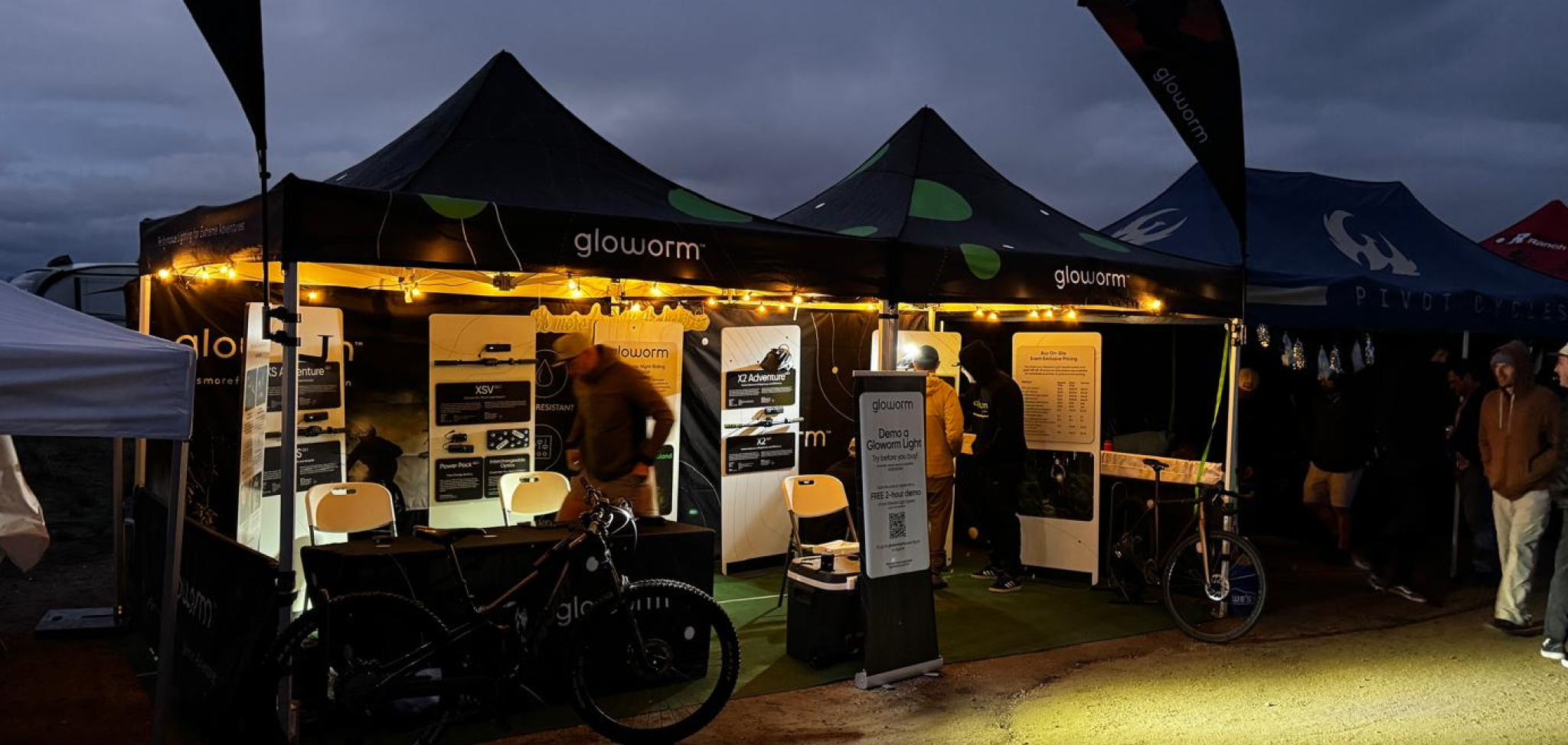 Gloworm Lights booth at night at the 24 Hours at Old Pueblo 2026, with display panels and bikes lit up under the event tent.